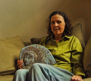 Smiling woman, sitting on couch, holding a bas-relief of an angel, created by her mother.