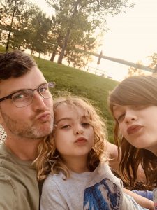 Man and two children blowing kisses at the camera, while sitting outside near a lake.