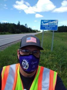 Man with mask on, standing roadside next to a sign that reads, Adopt a Highway, Sober Squad Bemidji, since 2019