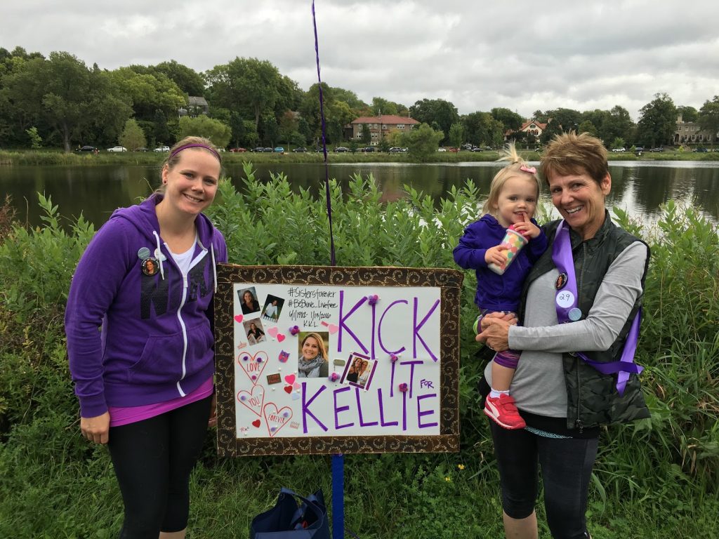 Two women, one holding a young girl, holding a sign that says Kick It for Kellie
